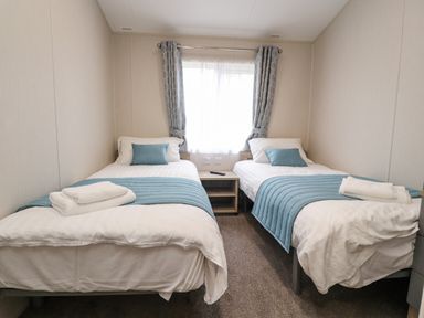 A bedroom with two single beds with blue throws and pillows a window with grey curtains and a bedside table at Fountain View in Warton near Carnforth