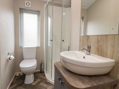 A bathroom with a white toilet beside a window with blinds and a corner shower next to a wooden countertop with a white sink and mirror at Fountain View in Warton near Carnforth