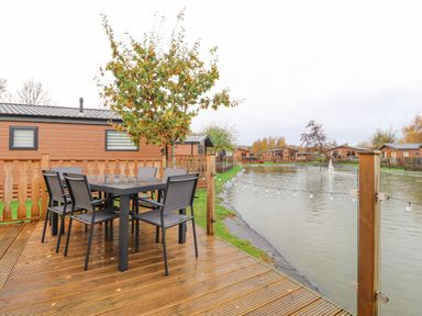An outdoor wooden deck with black dining table and chairs overlooking a pond with cabins around it at Fountain View in Warton near Carnforth