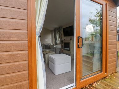 A view through an open sliding glass door into a living room with a grey ottoman coffee table sofa fireplace and mounted tv at Fountain View in Warton near Carnforth
