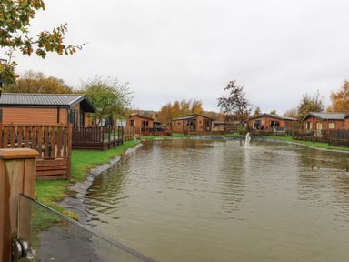 A pond with a fountain surrounded by wooden lodges and green grass at Fountain View in Warton near Carnforth