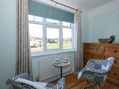 A room with two patterned chairs a small round table with mugs a wooden dresser and a window with curtains at Beck View in Sheringham