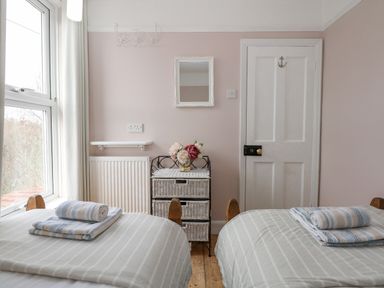 A bedroom with two single beds with folded towels on each bed a window with curtains a small chest of drawers with flowers and a door at Beck View in Sheringham