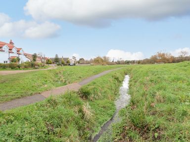 A narrow stream with grassy banks next to a path and houses in a rural area at Beck View in Sheringham