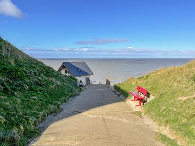 A path leading down to a small building by the sea with grass-covered hills on each side and a red bench on the right at Beck View in Sheringham