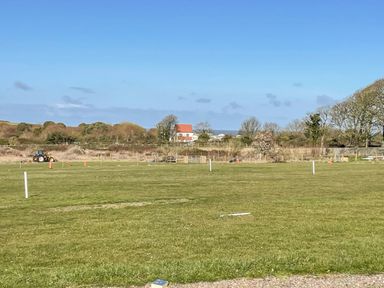 A grassy field with white markers and a tractor near a fenced area with trees and a house with a red roof in the background at Beck View in Sheringham