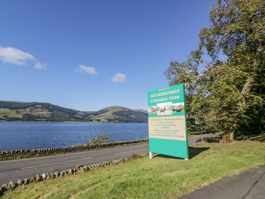 A road next to a body of water with a green sign for Auchengower Caravan Park and trees at 11 Mansion View in Helensburgh near Kilcreggan