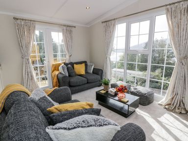 A living room with two grey sofas yellow cushions a black coffee table with a fruit bowl and a plant and large windows with curtains at 11 Mansion View in Helensburgh near Kilcreggan