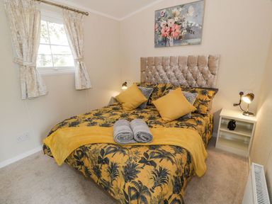 A bedroom with a bed with yellow and black floral bedding two rolled towels on the bed a window with curtains and a nightstand with a lamp and decoration at 11 Mansion View Helensburgh near Kilcreggan