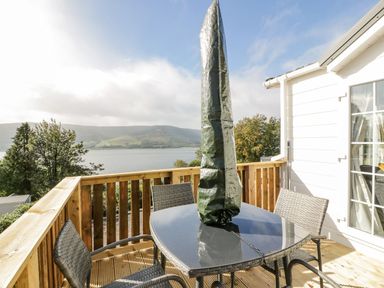 An outdoor wooden deck with a glass table and four wicker chairs overlooking a lake and hills at 11 Mansion View in Helensburgh near Kilcreggan