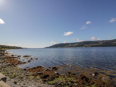 A rocky shoreline with seaweed along a calm body of water and hills in the distance at 11 Mansion View in Helensburgh near Kilcreggan