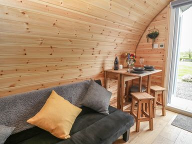 A wooden interior with a black couch with cushions and a small dining table with stools beside a door open to the outside at Parys Pod in Amlwch