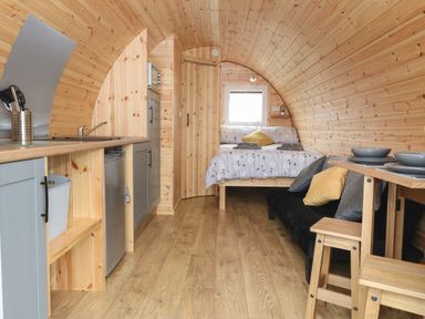 A curved wooden interior with a kitchen area including a sink and refrigerator a bed with floral bedding a small dining table with stools and a black sofa with pillows at Parys Pod in Amlwch