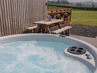 An outdoor hot tub with bubbling water next to a wooden picnic table and a fence with planters at Parys Pod in Amlwch