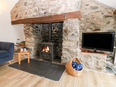 A living room with a stone fireplace with a wood stove a blue chair a small wooden table with teacups a flat screen TV and a basket with a blanket at Y Bwthyn in Dyffryn Ardudwy