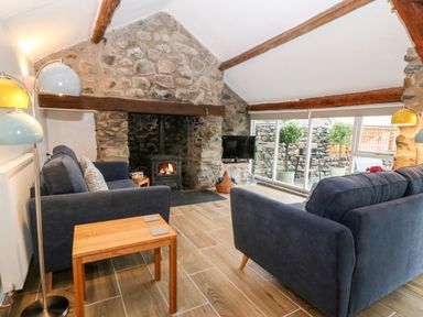 A living room with two blue sofas a stone fireplace a wood burning stove wood paneled floor and glass doors leading outside at Y Bwthyn in Dyffryn Ardudwy