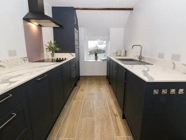 A kitchen with dark cabinets white marble countertops a window at the end and wood tile flooring at Y Bwthyn in Dyffryn Ardudwy