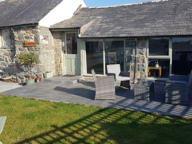 An outdoor patio with wicker chairs and a glass table outside a stone house with a green door at Y Bwthyn in Dyffryn Ardudwy