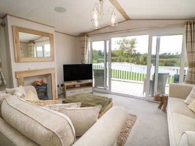 A living room with beige sofas a television a fireplace and glass sliding doors leading to a deck overlooking a pond at 10 Cheviot View in Haggerston near Lowick