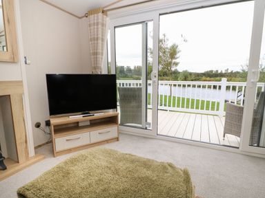 A living room with a television on a wooden stand and glass sliding doors opening to a balcony overlooking water at 10 Cheviot View Haggerston near Lowick