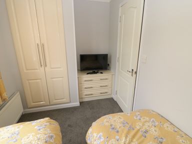 A bedroom with two beds covered in yellow floral bedspreads a cream wardrobe a chest of drawers with a television on top and a white door at Mildred's Lodge in Felton