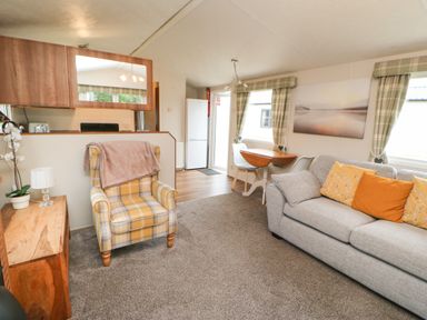 A living room with a gray sofa with cushions a yellow plaid armchair a wooden side table a small dining table with chairs and kitchen area at Hedgehog Retreat in Felton