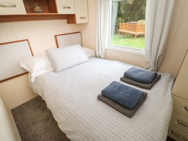 A bedroom with a bed made with striped bedding two folded towels on the bed and a window with curtains overlooking a garden at Hedgehog Retreat in Felton