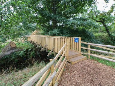A wooden footbridge over a small stream with safety railings and a caution sign surrounded by trees at Hedgehog Retreat in Felton
