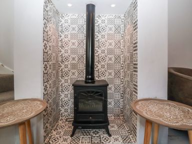 A wood burning stove with a black pipe surrounded by patterned tiles between two small round tables at Secret Cottage Southwold