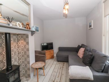 A living room with a gray sectional sofa a small wooden side table and a stove with patterned tiles at Secret Cottage Southwold