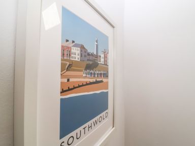 A framed picture of Southwold showing buildings a lighthouse beach huts and the sea on a white wall at Secret Cottage Southwold