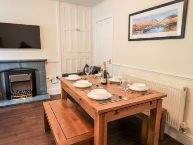 A dining area with a wooden table set with plates glasses and a bottle a bench a fireplace and a mounted television at 6 Calgarth View in Windermere