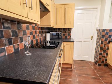 A kitchen with wooden cabinets a black countertop two wine glasses a stove a microwave and tiled walls at 6 Calgarth View in Windermere