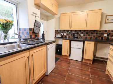 A kitchen with wooden cabinets a sink a dishwasher a small fridge and flowers by the window at 6 Calgarth View in Windermere