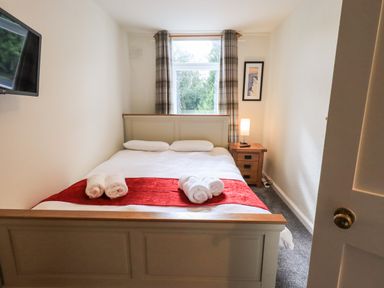 A bedroom with a bed with white and red bedding a nightstand with a lamp a window with curtains and a wall-mounted tv at 6 Calgarth View in Windermere