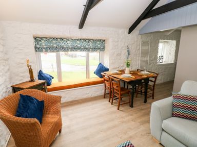 A dining area with a wooden table set for four next to a window seat and wicker chair at Bluebell Cottage in Jameston near Manorbier