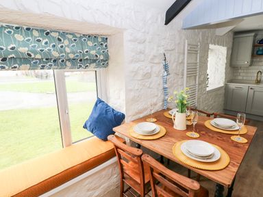 A dining area with a wooden table set for four next to a window with a floral Roman blind at Bluebell Cottage in Jameston near Manorbier