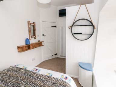 A bedroom corner with a bed partly visible a white wooden door a wall shelf with a blue vase a round hanging shelf and a laundry basket at Bluebell Cottage in Jameston near Manorbier