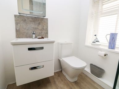 A bathroom with a white sink cabinet and toilet next to a window with blinds and a striped jug at Bluebell Cottage Jameston near Manorbier