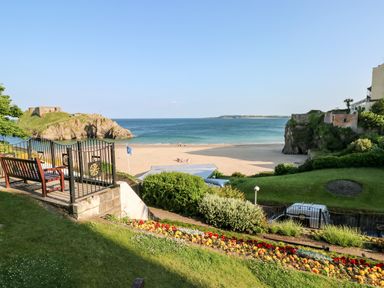 A garden with flowers, benches, and a gate overlooking a sandy beach and sea at Bluebell Cottage in Jameston near Manorbier