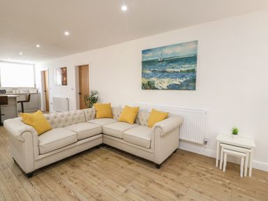 A living room with a beige sectional sofa with yellow cushions a painting of sailboats on the sea and nested white side tables at The Church in Holyhead
