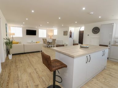 A modern open-plan living space with kitchen island, brown chairs, beige sofa, dining table, wall clock and TV at The Church in Holyhead