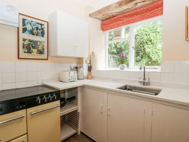 A kitchen with a stove a toaster and a kettle on the counter a sink under a window with a flower vase and a framed picture on the wall at Ham cottage East Lambrook near South Petherton