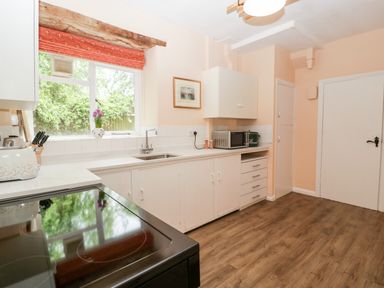 A kitchen with white cabinets a window with a red curtain and wooden flooring at Ham cottage East Lambrook near South Petherton
