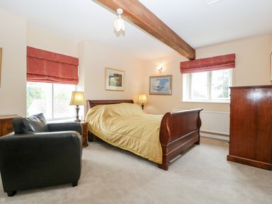 A bedroom with a wooden bed covered with a yellow quilt a black armchair two windows with red blinds two bedside tables with lamps and a wooden cabinet at Ham cottage in East Lambrook near South Petherton