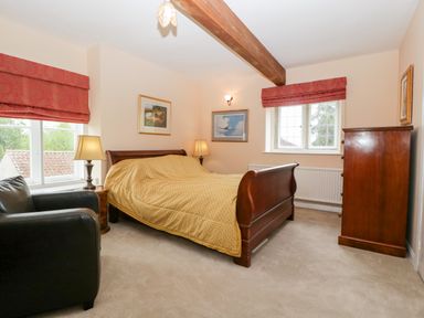 A bedroom with a wooden bed a black armchair two windows and a wooden chest of drawers at Ham cottage East Lambrook near South Petherton