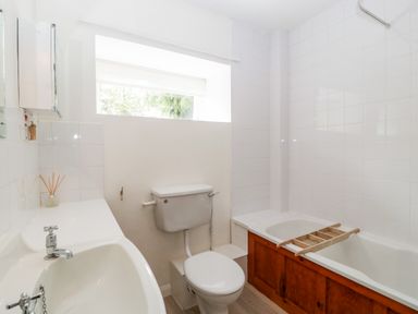 A bathroom with a white sink, toilet, bathtub with a wooden panel, and a window above the toilet at Ham cottage in East Lambrook near South Petherton