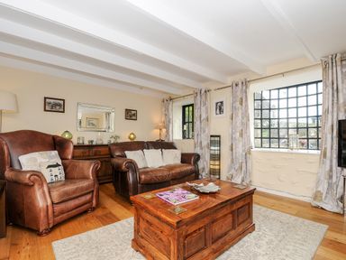 A living room with brown leather armchair and sofa a wooden chest coffee table windows with curtains and wall mirror at Church View Cottage Near Looe