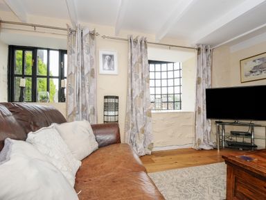 A living room with a brown leather sofa white pillows patterned curtains a glass window a flat screen tv on a stand and a wooden coffee table at Church View Cottage Near Looe
