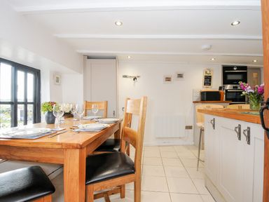 A dining area with wooden table set with plates and glasses next to a kitchen with white cabinets and appliances at Church View Cottage Near Looe